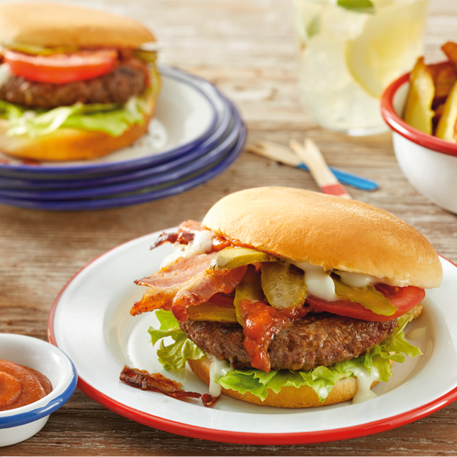 Kaksi hampurilaista, joissa on pekonia, suolakurkkuja ja tomaattia, sekä ranskalaisia ja juoma.