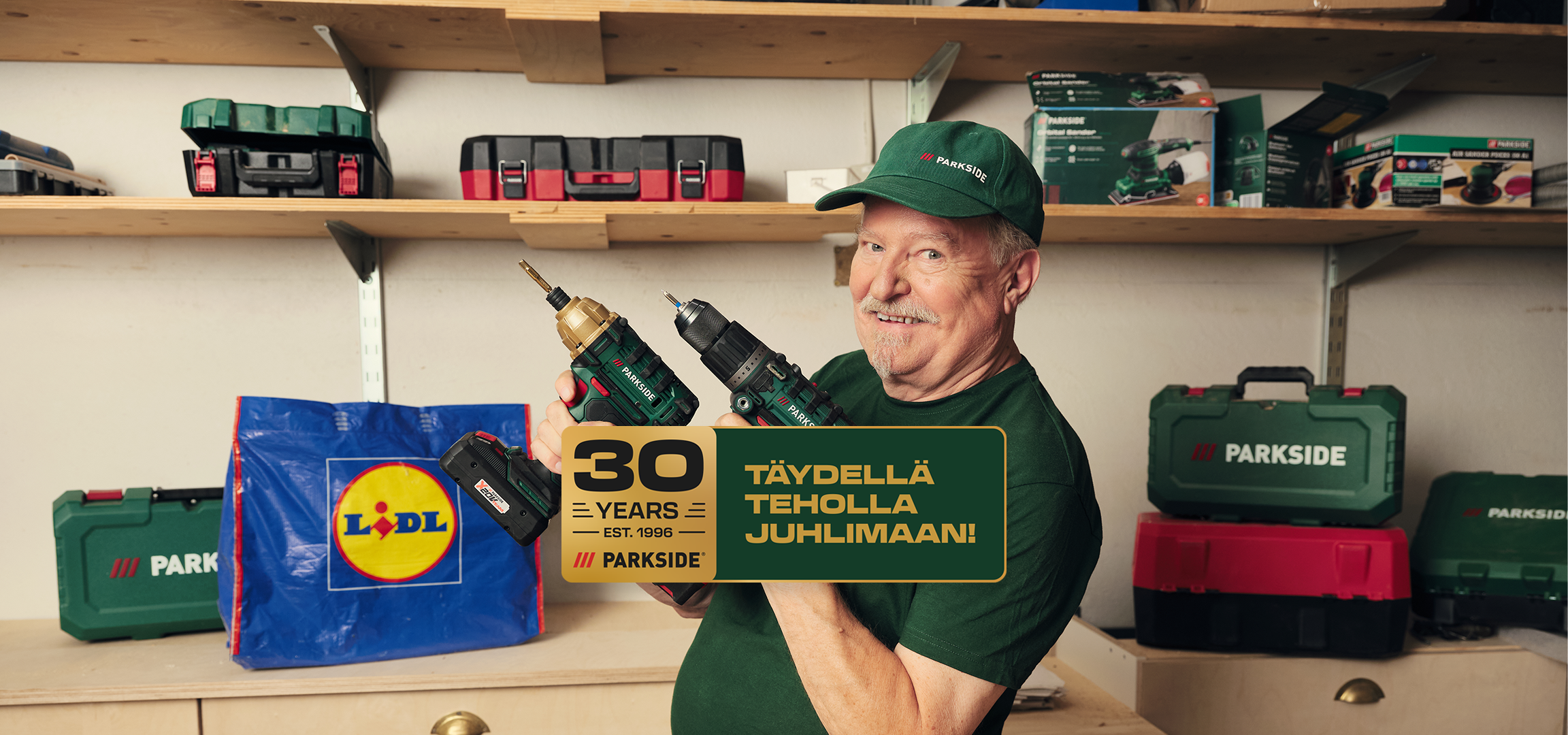 Mies hymyilee ja pitelee kahta akkuporakonetta, taustalla työkaluja ja ostoskassi. Teksti: 30 vuotta, täydellä teholla juhlimaan!