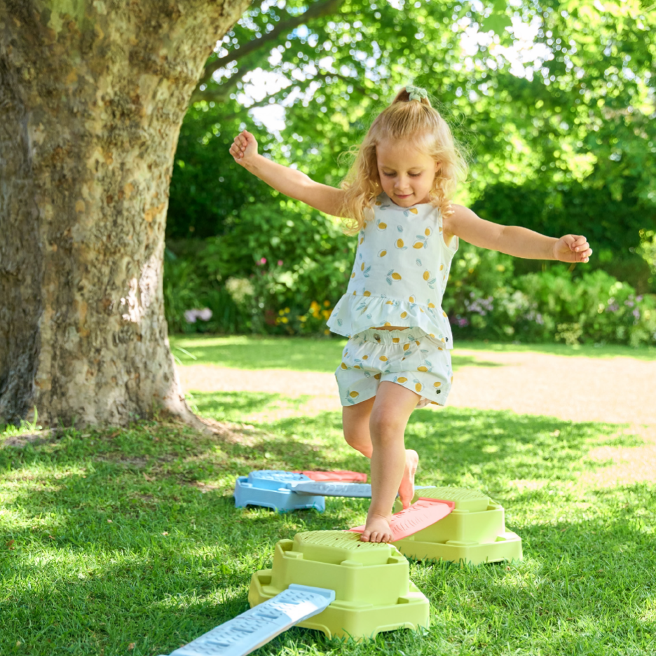 Pieni lapsi tasapainoilee värikkäillä tasapainolaudoilla nurmikolla puun alla.